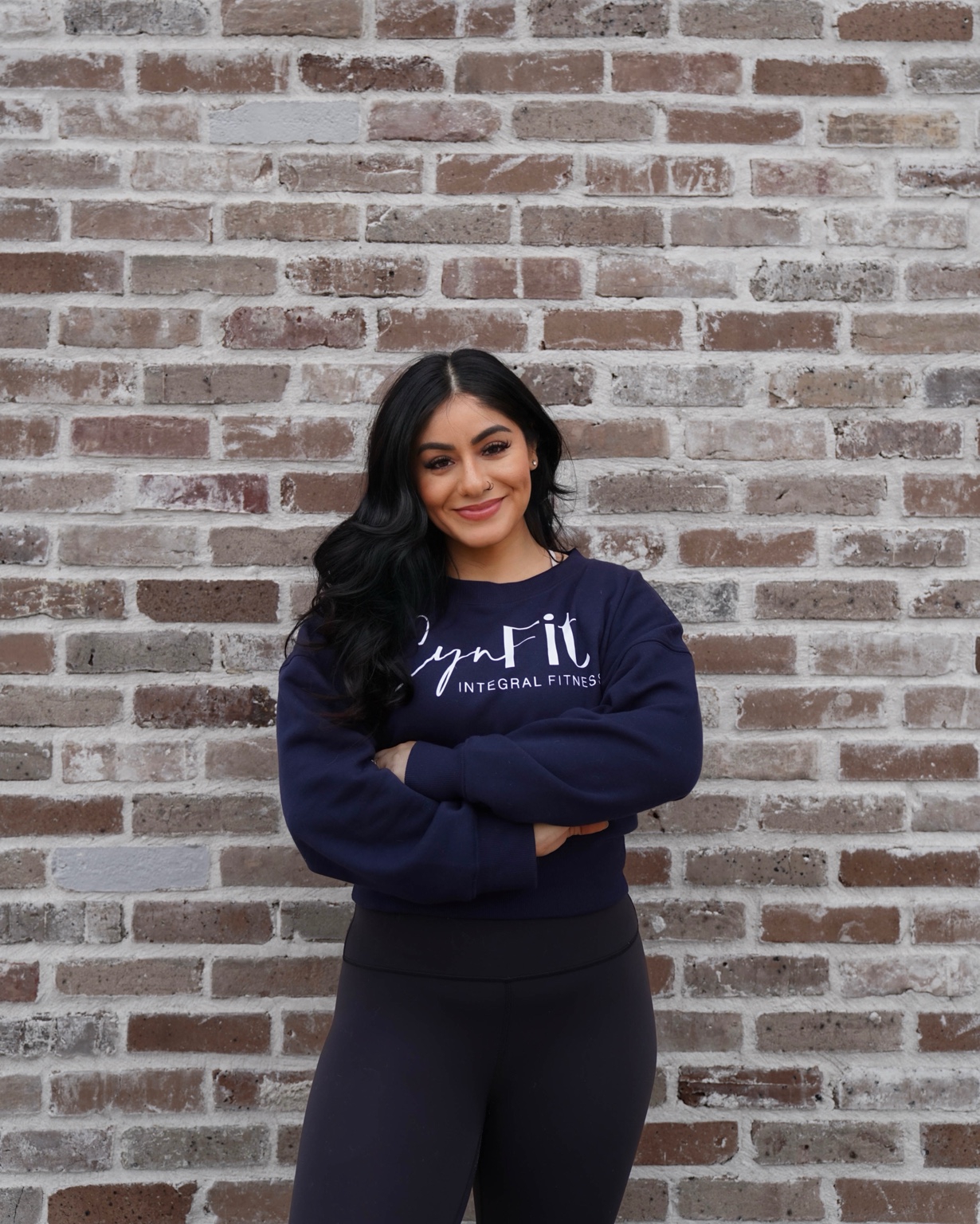 Coach Cynthia Martinez smiling in a navy CynFit sweatshirt with her arms crossed in front of a brick wall